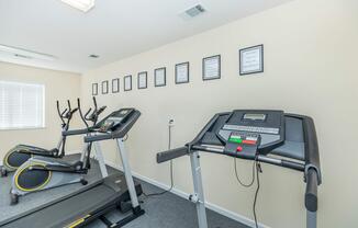A small gym area featuring two treadmills and an elliptical machine. The wall is adorned with framed motivational quotes. The flooring is carpeted, and there is natural light coming from a window.