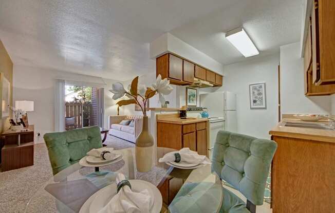 A bright dining and kitchen space here at Northridge Court showcasing warm wood cabinetry, light countertops, and a glass dining table with upholstered chairs, opening into a comfortable living area with soft carpeting, neutral walls, and a sliding glass door that brings in natural light.