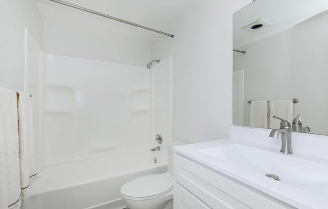 A clean, modern bathroom featuring a white bathtub with a shower, a white toilet, and a sleek vanity with a sink. The walls are painted in a light color, and there is a large mirror above the sink. Towels are neatly hung on a rack, contributing to the tidy and airy atmosphere of the space.
