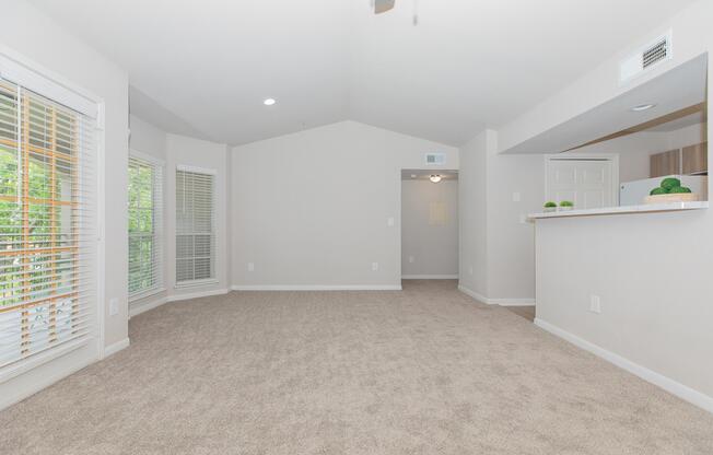 Spacious, light-filled living area with beige carpet and neutral wall color. Features large windows with blinds, a ceiling fan, and a kitchen bar area. Open layout leading to a hallway, creating a welcoming atmosphere suitable for various uses.