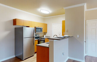 A compact kitchen here at Landing at Mansfield with warm wood cabinets, stainless steel fridge and range, raised breakfast bar with place settings, and soft overhead lighting in a clean, bright layout.