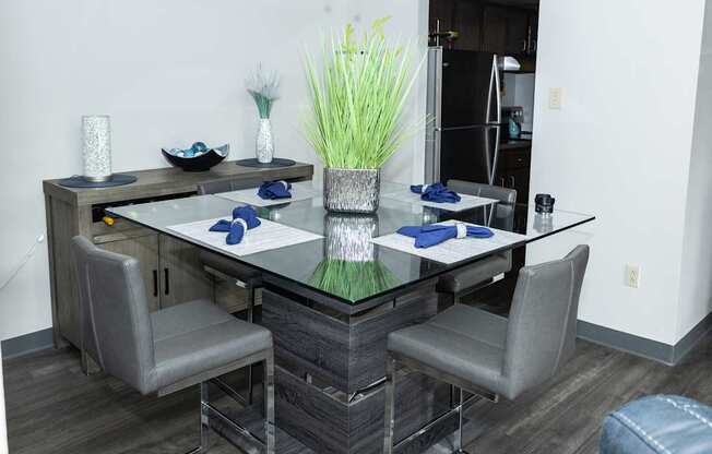 A glass dining table with four grey chairs and a vase of grass in the middle.
