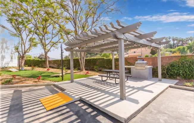 One-Bedroom Apartments In Colton, CA - Ardella At Reche Canyon - Picnic Area Under Pergola With Wheelchair Ramp, BBQ Grill, Table With Benches, Beside Cornhole Area.