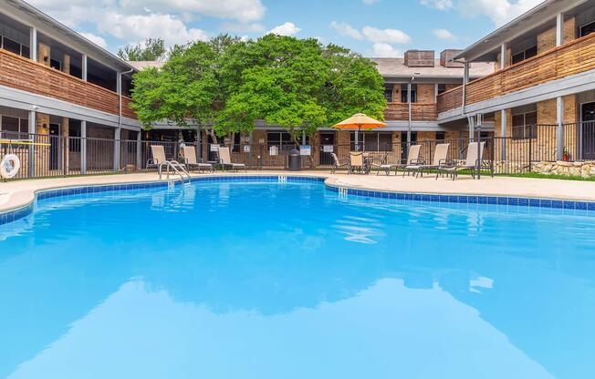 A bright, inviting swimming pool surrounded by lounge chairs and an orange umbrella, set within a courtyard of a two-story building. Lush greenery and a clear blue sky create a relaxed atmosphere, perfect for relaxation and leisure.