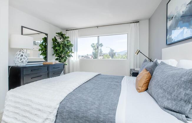 Bright and modern bedroom featuring a large bed with gray bedding and decorative pillows. A dark dresser is styled with a contemporary lamp and a potted plant. Sunlight streams through a large window, enhancing the airy atmosphere. A framed artwork hangs on the wall, adding a touch of elegance.