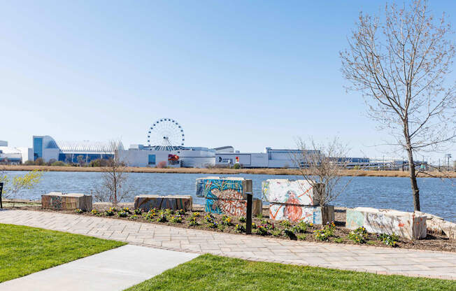 A park with a walkway, a tree, and a body of water with a building in the background.