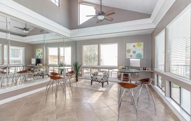 A modern dining area with a round table and stylish high chairs, surrounded by large windows with blinds. The walls are painted a muted gray, and there's a mirrored wall enhancing the space. A colorful abstract art piece is on display, and greenery adds a touch of nature to the stylish interior.