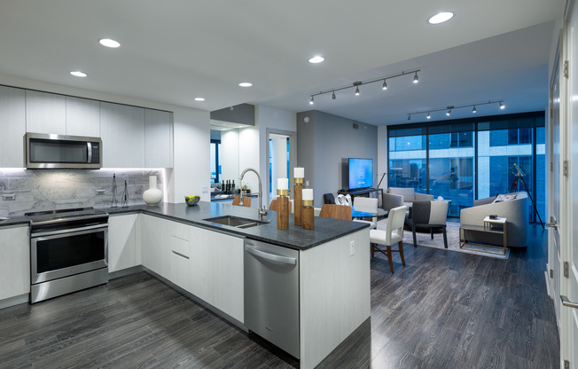 Open-concept kitchen, living, and dining space with dark wood-style floors, white handleless cabinets, stainless appliances, full-width floor-to-ceiling windows, and a gray accent wall.