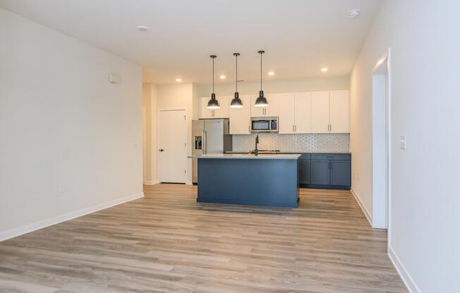 A modern open-concept kitchen and living area featuring light wood flooring, dark cabinets, and stainless steel appliances. The kitchen island has pendant lighting above, and there's a tile backsplash. The space is well-lit with natural light and has a minimalist design.