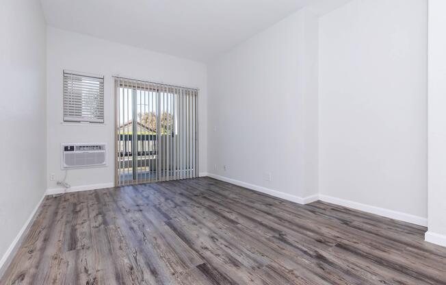 Empty room with light gray walls and wooden flooring. There are vertical blinds covering a large window that opens to a balcony. An air conditioning unit is mounted on the wall near the window. The space is bright and airy, with ample natural light coming in.