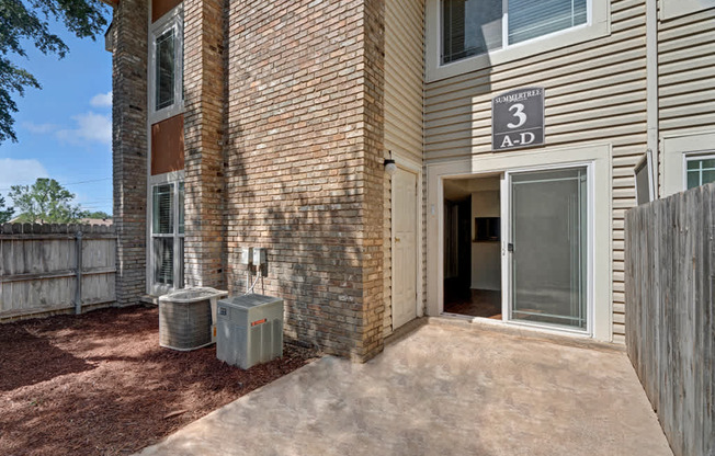 A private ground-level patio here at Summertree Place featuring a concrete patio surface, a sliding glass door leading into the apartment, brick and siding exterior walls, landscaped mulch along the building, and a fenced boundary that creates a defined outdoor space with convenient access to the interior.