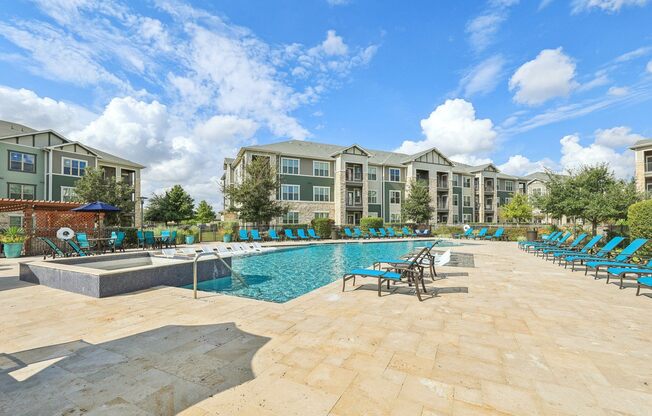 A large swimming pool surrounded by lounge chairs in front of apartment buildings.