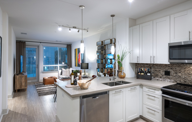 Open-concept kitchen, dining, and living space with white walls, light wood-style floors, white cabinets, stainless steel appliances, and a large window and glass door leading from the living area to the balcony.