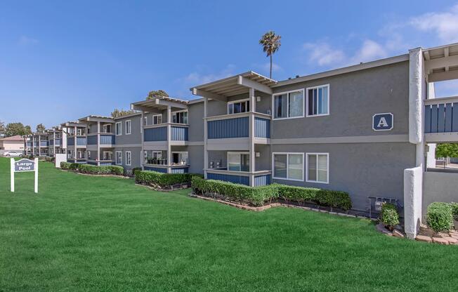 A row of modern apartment buildings with balconies, set on a well-maintained grassy area. The structures have gray exteriors and blue accents. A palm tree is visible beside the buildings. A sign labeled "Lawn Post" stands in the foreground, indicating the layout of the property. Bright blue sky completes the scene.