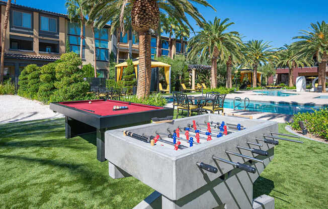 A red table tennis table with blue paddles and red balls is in the foreground of a pool and palm trees.