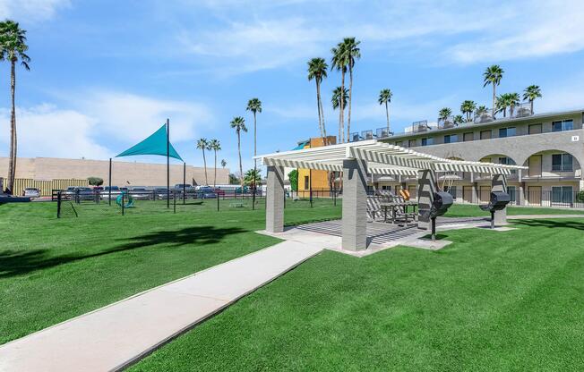 A well-maintained outdoor area featuring a grassy lawn, a pergola with picnic tables, and BBQ grills. In the background, there are palm trees and an apartment building, along with a blue shade structure. The sky is clear and bright, creating a welcoming atmosphere for outdoor activities.