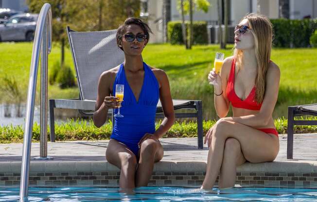 Two women sitting by a pool, one in a blue swimsuit and the other in a red swimsuit.