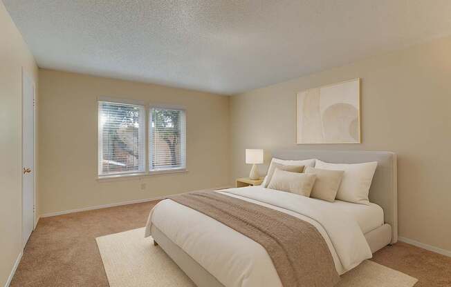 A bright bedroom here at Summertree Place featuring a neatly made bed with neutral bedding, a soft area rug over carpeted floors, a bedside table with a lamp, and large windows that bring in natural light, creating a calm and comfortable retreat.