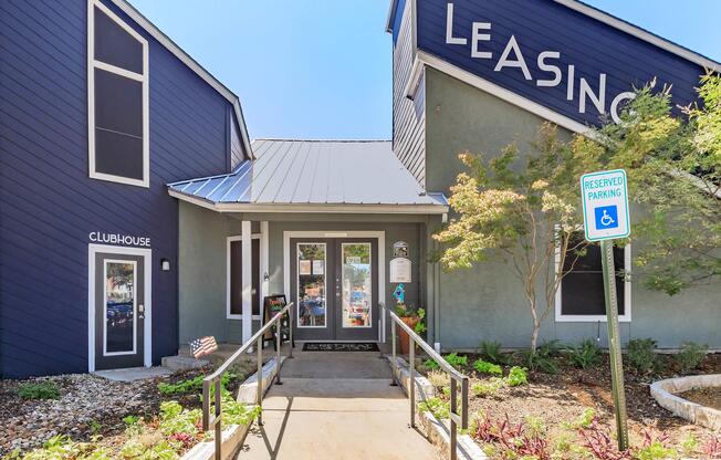 A modern leasing office with a blue and grey exterior. The entrance features a ramp, signage reading "LEASING" and "CLUBHOUSE," and manicured landscaping with plants. A reserved parking sign is visible nearby. The building has large windows that provide a welcoming atmosphere.