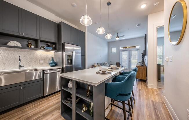 A kitchen with an island, a large farmhouse-style sink, stainless steel appliances, and pendant lights near a living room and bedroom.