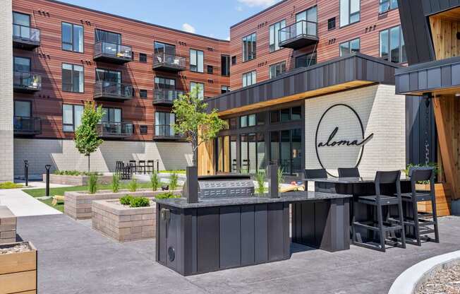 A modern outdoor seating area with a bar and chairs is situated in front of a building with a sign that reads "Alma.".