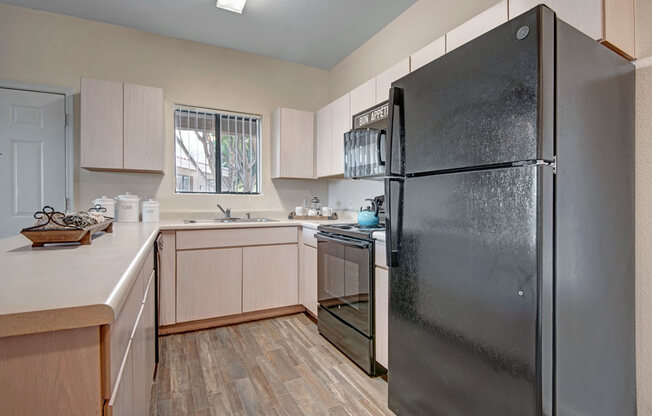 A bright and inviting kitchen here at Pala Mesa featuring light wood cabinetry, expansive counter space, and a double-basin sink positioned beneath a sunlit window. A full appliance package complements the layout, while wood-style flooring and overhead lighting enhance the clean, functional design and open feel of the space.