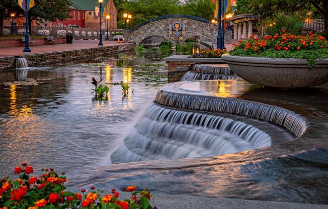 a fountain in a city park with flowers and a bridge