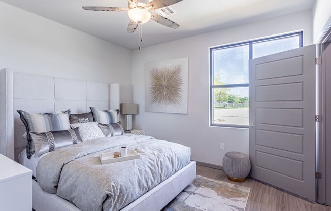 Bright bedroom here at 49 West Apartments with a large bed featuring gray and silver pillows and comforter, modern art on the wall, a ceiling fan, and a sunlit window. Cozy and serene atmosphere.