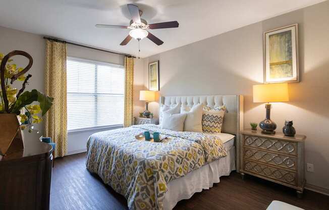 A bedroom with a bed, nightstand, and a ceiling fan in the 826 square foot 1 bedroom, 1 bathroom apartment at The Brownstones Townhome Apartments in the Far North Dallas neighborhood of Dallas, TX.
