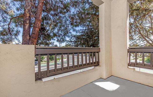 View from a balcony displaying a corner perspective with a concrete floor and beige walls. A wooden railing separates the balcony from an outdoor scene featuring trees and a street visible in the background under a clear sky. Natural light casts shadows on the floor.