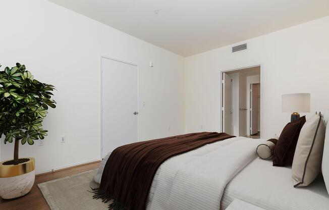 A cozy bedroom featuring a neatly made bed with brown and white bedding, a bedside lamp, and a decorative plant in a gold pot. The room has light-colored walls and wooden flooring, with a door visible in the background leading to another area.