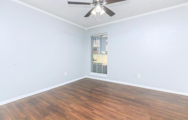 Empty room with light gray walls and hardwood flooring. A ceiling fan with lights is mounted in the center. There is a window with horizontal blinds on the right side, allowing natural light to enter. The room has a simple, clean appearance, suitable for various purposes.