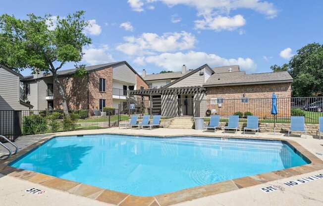 A swimming pool surrounded by chairs and a fence with the leasing office in the background at The Oaks of Denton Apartments in Denton, TX