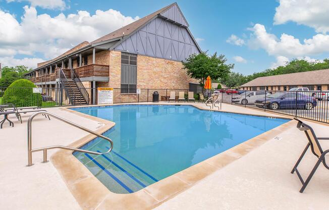 A clear swimming pool surrounded by lounge chairs and an orange umbrella, located next to a multi-story brick building. Lush greenery is visible, and parked cars are in the background under a bright blue sky with fluffy white clouds.