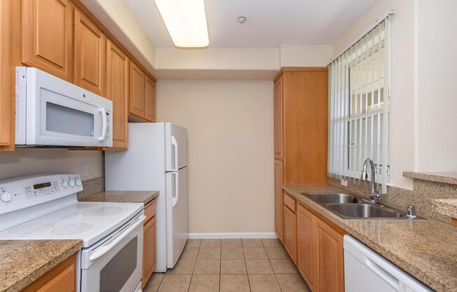 A modern kitchen featuring light wood cabinetry, a white microwave and refrigerator, an electric stove with an oven, a double sink with a sleek faucet, and granite countertops. The space has beige walls and tile flooring, with natural light coming through a window.