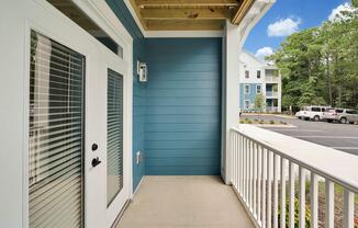 A modern porch area featuring a blue accent wall, white door with vertical blinds, and a railing. In the background, there are parked cars and an apartment building surrounded by greenery under a clear sky.