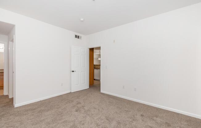 A spacious, empty room with light beige carpet and white walls. A door leads to another room, and there is visible natural light. No furniture or decorations are present, providing a blank canvas for potential decoration.