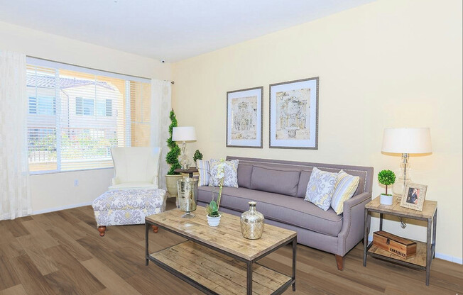 Bright living room with light-colored walls, featuring a gray sofa adorned with decorative pillows, a patterned armchair, and a wooden coffee table. Two framed artworks hang above the sofa, and a small side table with a lamp is nearby. Natural light streams through a large window, illuminating the space.