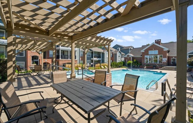 A wooden pergola over a table with chairs and a pool in the background.