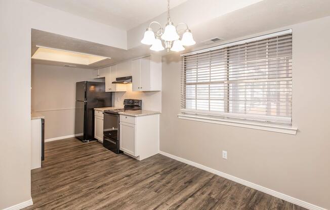 A modern kitchen with a refrigerator, stove, and white cabinetry. The walls are a neutral color, and there's a window with blinds letting in natural light. The flooring is a wood-like laminate, and a light fixture hangs from the ceiling. The space appears clean and well-organized.