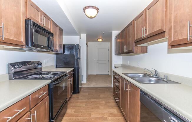 Modern kitchen featuring dark wood cabinets, stainless steel appliances including a microwave and oven, and a double sink. The countertop is light-colored, and the floor is a warm wood tone. There is an entrance visible at the far end of the room, along with a black refrigerator.