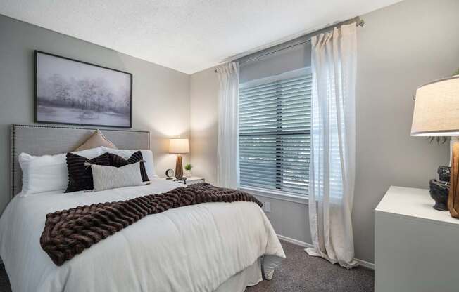 A bedroom with a large bed and a window with white curtains.