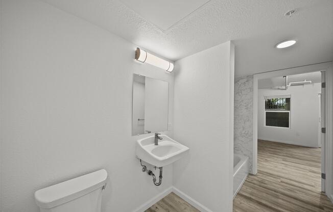 A clean, modern bathroom featuring a white sink with a mirror above it, a white toilet, and light wood flooring. The walls are painted white, and there is a marble accent wall visible. A doorway leads to another room with a window, providing natural light.