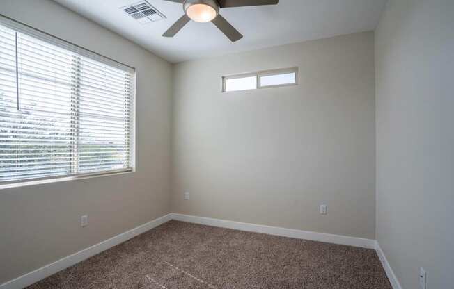 A carpeted bedroom has a window and ceiling fan in an apartment at Village Greens of Queen Creek