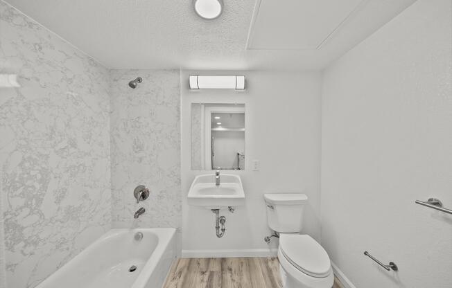A modern bathroom featuring a white bathtub with a marble surround, a wall-mounted sink with a mirror above it, and a toilet. The walls are mostly white, and the flooring is light wood. A ceiling light fixture illuminates the space, creating a bright and clean ambiance.