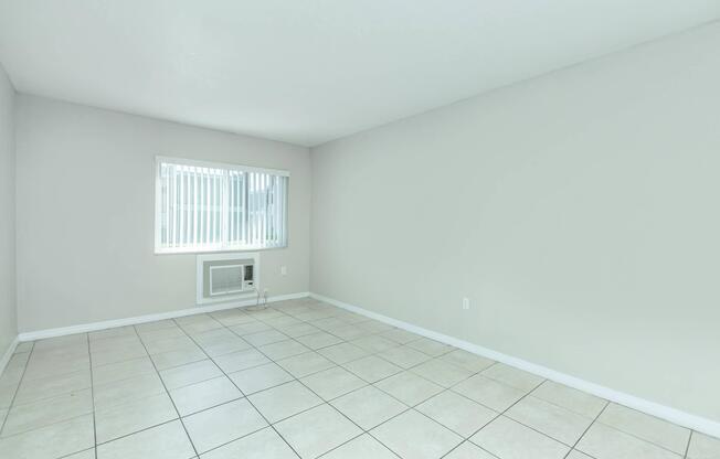 A vacant room with light gray walls and tiled flooring. There is a window with white blinds letting in natural light, and an air conditioning unit beneath it. The space is empty, creating a clean and minimalistic atmosphere.
