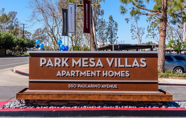 A brown sign for Park Mesa Villas Apartment Homes sits in front of a tree.