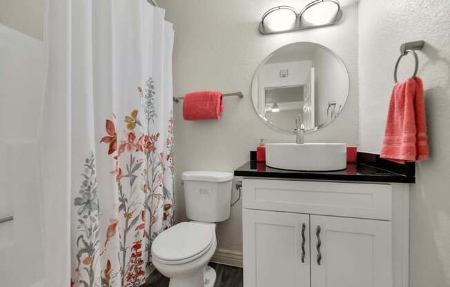 A white toilet with a floral shower curtain and a pink towel hangs on a wall next to a round mirror at Urban WalkApartments on 6th Avenue, Arizona 85016