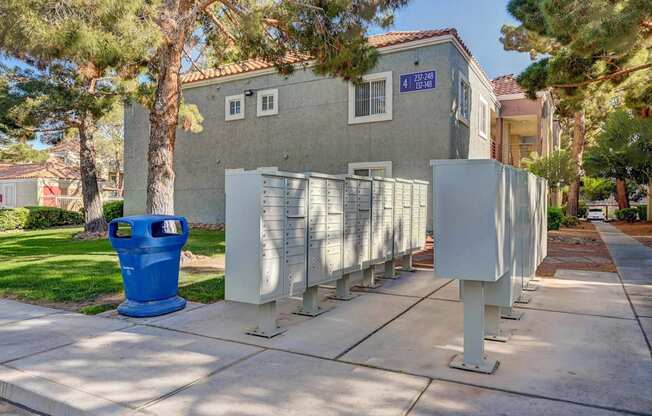A blue trash can is in front of a grey building with a tree in front of it.
