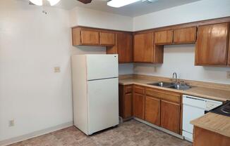 A small kitchen space featuring light wood cabinetry, a white refrigerator, double sink, and a dishwasher. The walls are painted a light color, and the flooring has a tile pattern. There is a ceiling fan and overhead lighting, creating a bright and functional environment.